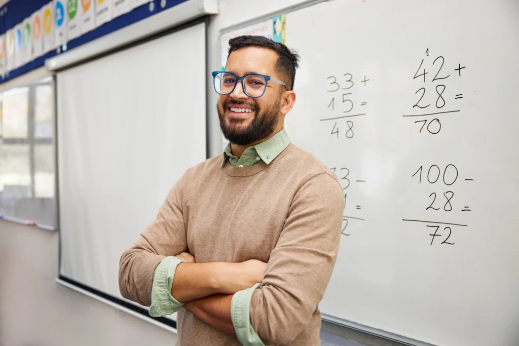 Male maths teacher with arms folded smiling in front of a whiteboard showing handwritten addition sums.