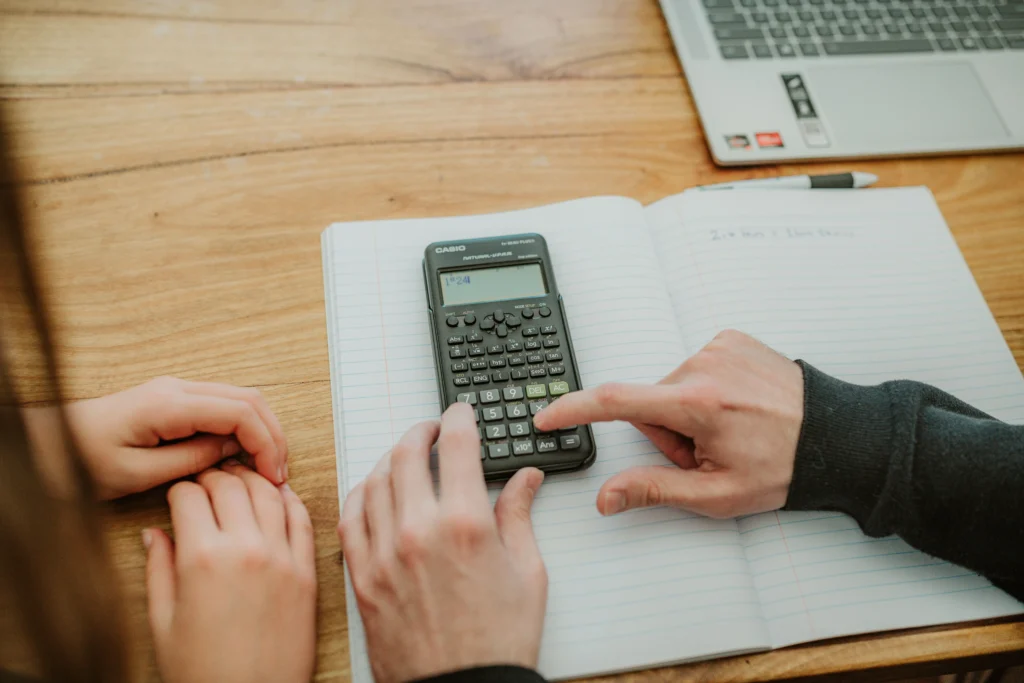 Close-up of a student's and tutor's hands using a scientific calculator alongside handwritten maths working in a lined notebook.