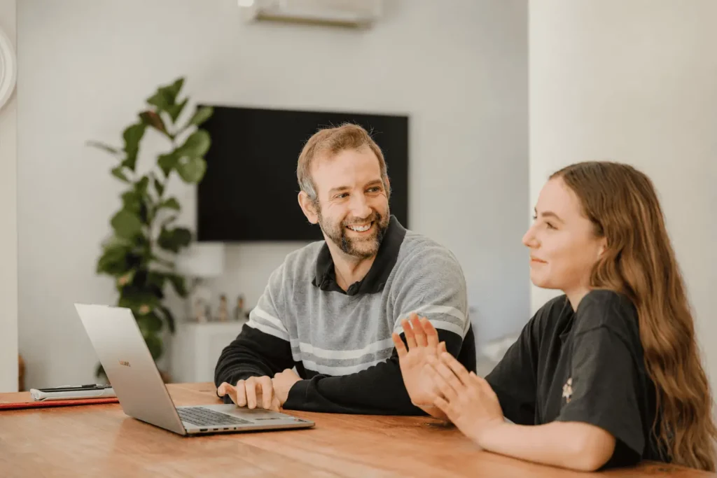 Male maths tutor smiling while discussing a problem with a female secondary student during an online tutoring session at a home desk in Perth.