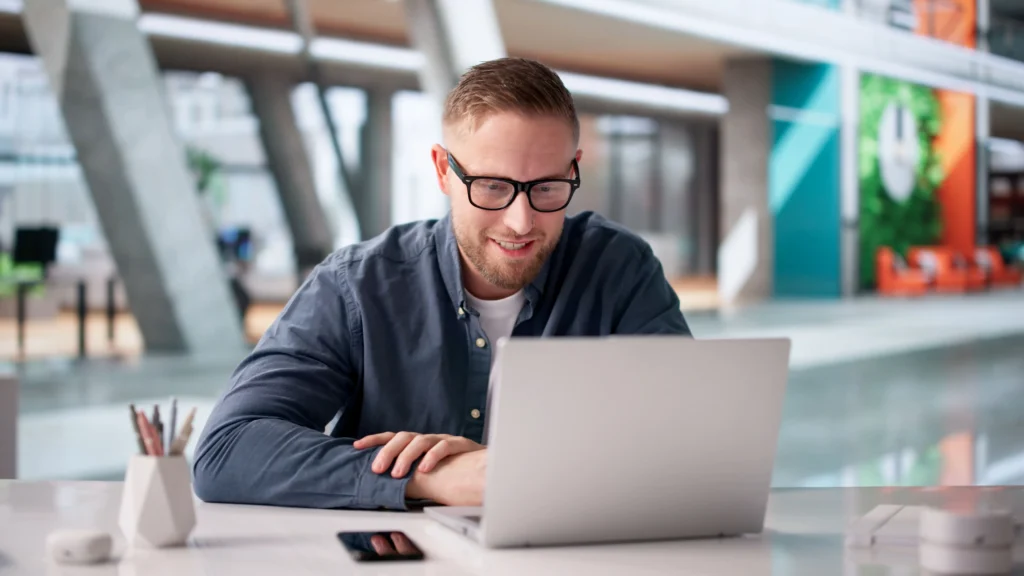 Male tutor wearing glasses working on a laptop at a desk while preparing online maths lessons.