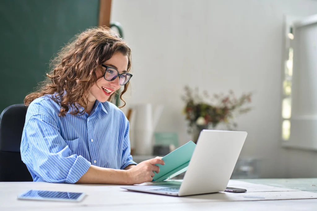 Woman wearing glasses smiling while working on a laptop with a notebook open beside her.