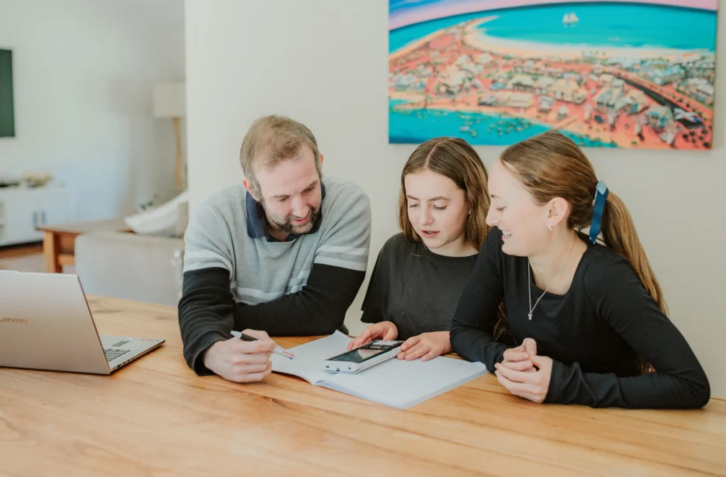 Maths tutor pointing at a workbook with a pen while two students follow along with a calculator during a tutoring lesson.