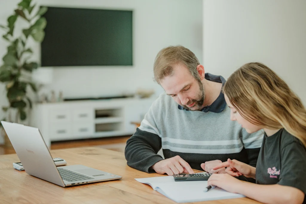 Maths tutor and a female secondary student concentrating on a calculator and workbook during an online tutoring session.