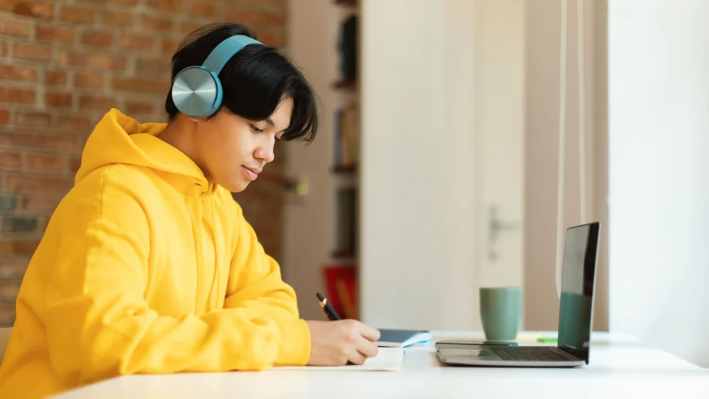 Secondary student wearing headphones writing in a notebook beside a laptop during an online study session at home.